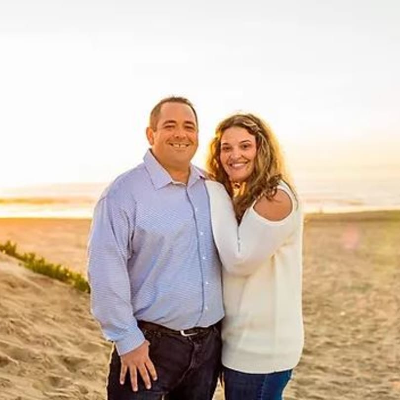Smiling couple standing on a sandy beach with the ocean and sunset in the background.