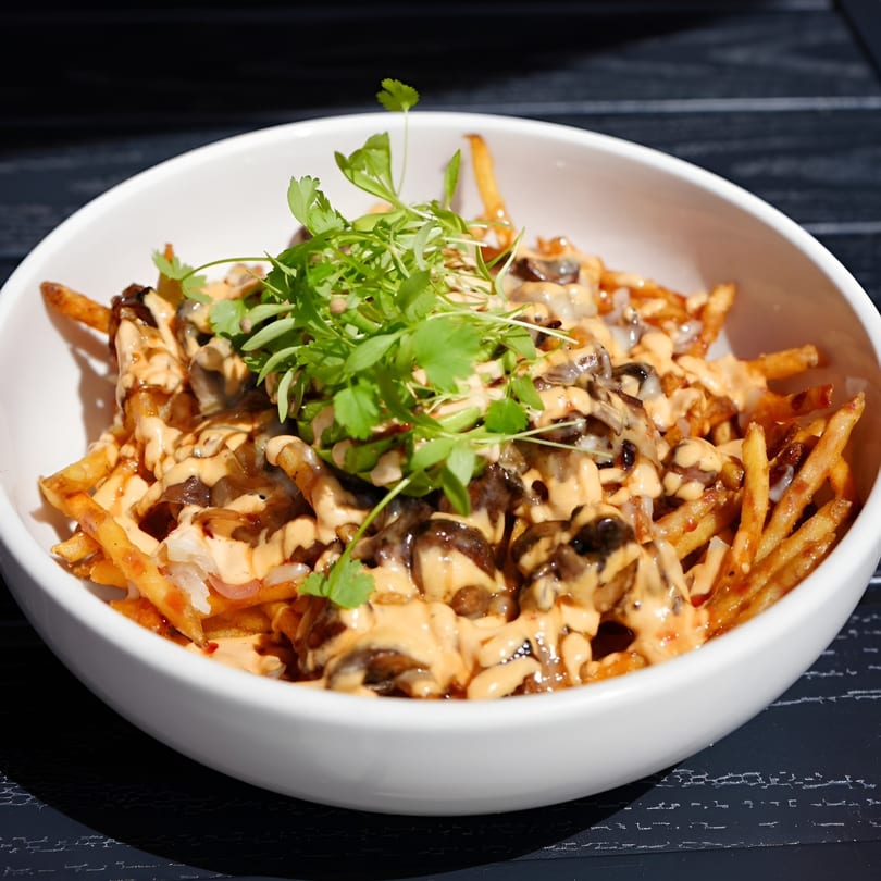 A white bowl of golden french fries topped with mushrooms, sauce, and fresh herbs.