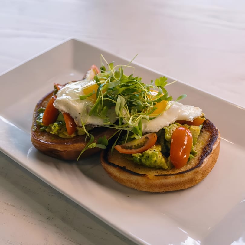 Avocado toast with fried eggs, cherry tomatoes, and microgreens on a white plate.