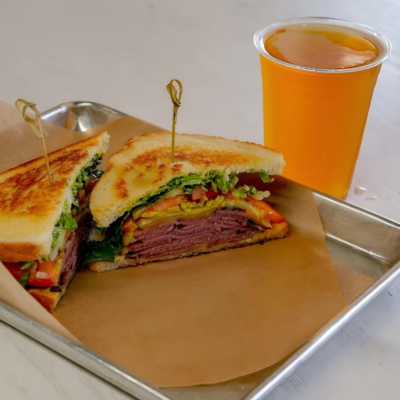 A sliced pastrami sandwich with lettuce, tomato, and pickles on toasted bread, served with an orange drink.