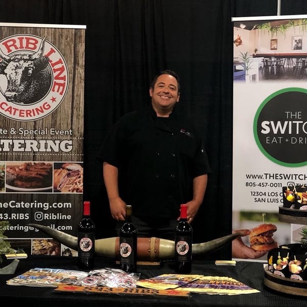 A smiling chef stands behind a table with Ribline Catering and The Switch SLO banners.