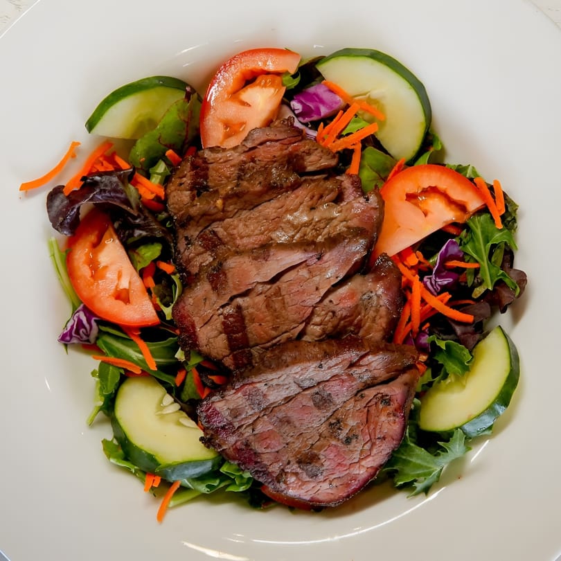 Grilled steak slices on a fresh green salad with tomatoes, cucumbers, and shredded carrots.