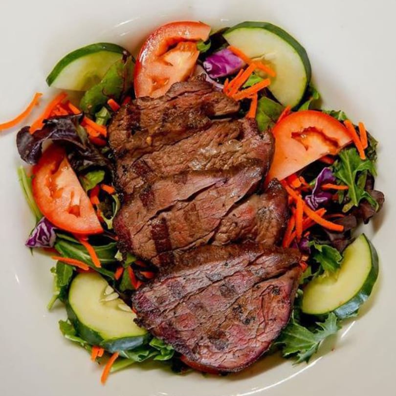 Grilled steak slices on a bed of mixed greens with tomatoes, cucumbers, and shredded carrots.
